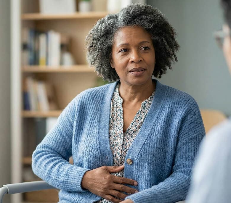 A woman in a blue cardigan sits and talks with another person in an office setting, with shelves in the background.